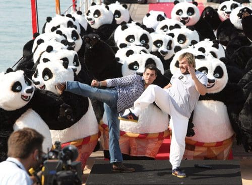Two men striking playful martial arts poses on a stage, surrounded by numerous large panda mascots. The scene takes place outdoors near water on a sunny day, creating a lively and whimsical atmosphere.