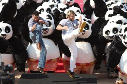 Two men dressed casually, one in a checkered shirt and the other in a gray t-shirt, perform expressive kicks on a stage. Behind them, a crowd of large panda costumes adds a playful atmosphere to the event.