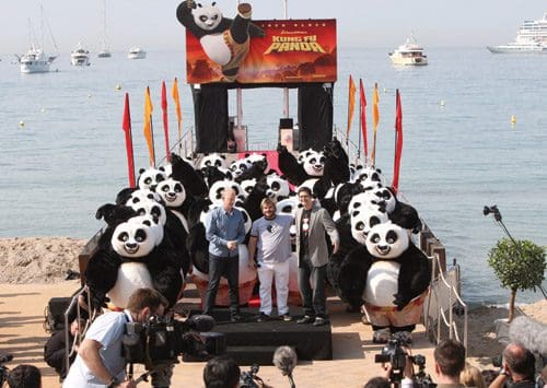A promotional event for 'Kung Fu Panda' featuring several people on a stage surrounded by performers in panda costumes, with a coastal backdrop and boats in the water.