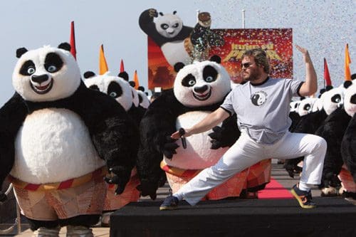 A man performing a martial arts pose on a stage, surrounded by several large panda characters in costumes during a colorful event. A festive atmosphere is enhanced by confetti and decorations in the background.