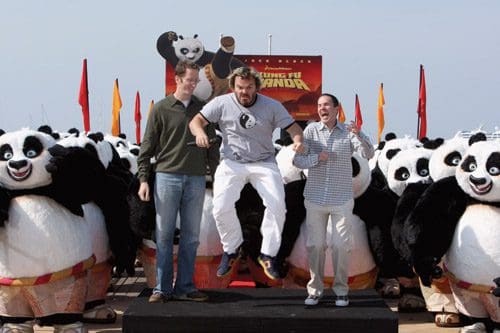 Three men on a platform are celebrating in front of a large group of panda mascots at an outdoor event. One man is mid-jump, while the others smile. Colorful flags decorate the scene, emphasizing a festive atmosphere related to 'Kung Fu Panda'.