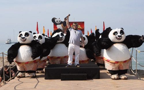 A man in casual clothing stands on a stage, raising a microphone, surrounded by several large panda costumes. The scene is set outdoors by the beach, with flags and a banner in the background.