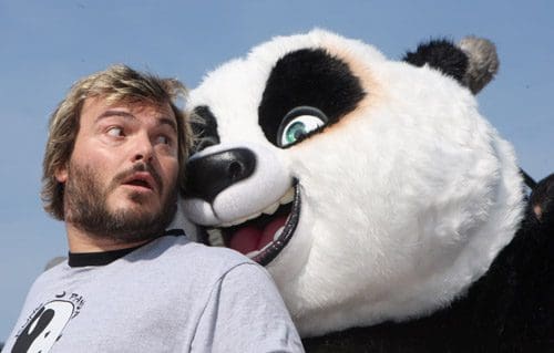 A man with messy hair in a gray t-shirt looks startled, glancing back at a large, friendly panda character beside him. The panda has a big smile and wide eyes, with a clear blue sky in the background.