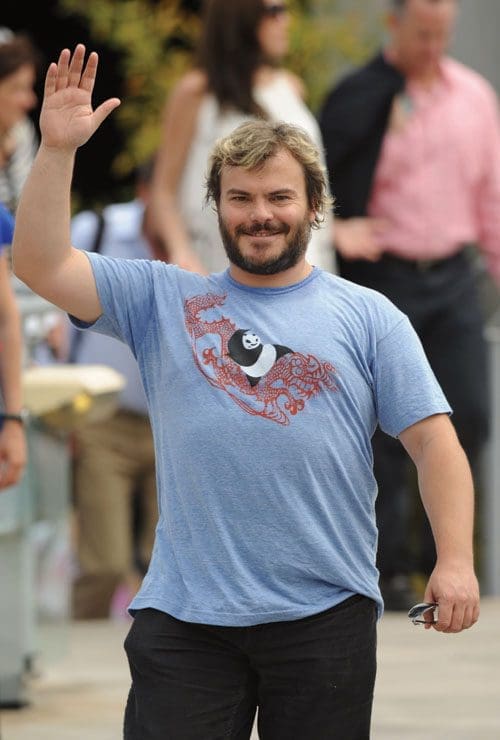 A smiling man with a beard waves while walking outdoors, dressed in a blue t-shirt featuring a graphic design, and black pants. In the background, people are casually socializing on a sunny day.