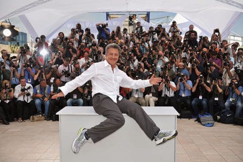A man in a white shirt and gray pants sits playfully on a table while posing for a large crowd of photographers at an outdoor event, with numerous camera flashes illuminating the scene.