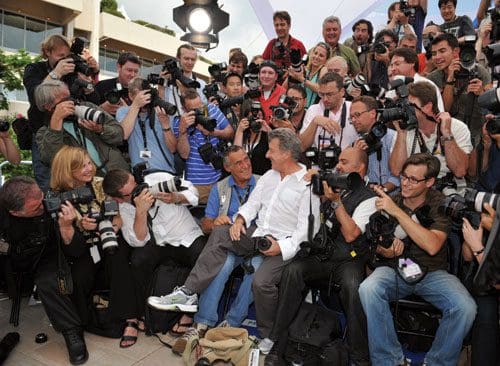 A crowded scene with numerous photographers gathered around a well-dressed man seated in the center. The group consists of various individuals equipped with professional cameras, some capturing the moment and others looking on.
