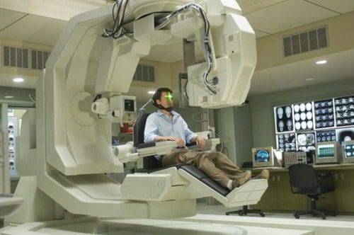 A man seated in a futuristic medical apparatus with a headset, in a clinical room filled with medical equipment and digital screens displaying scans.