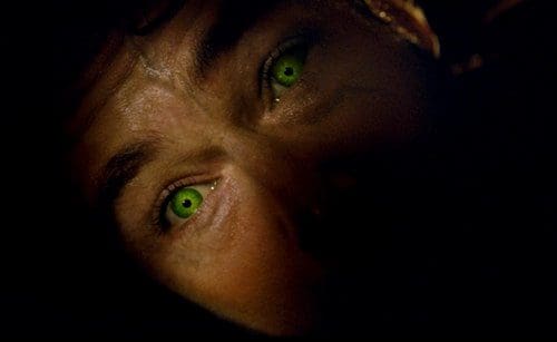 Close-up of a person's face with striking green eyes illuminated against a dark background, conveying a sense of intensity and intrigue.