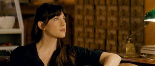A woman with long, dark hair and a serious expression sits on a sofa in a cozy room. She wears a black shirt and gazes thoughtfully into the distance, with a desk lamp casting a soft glow beside her.