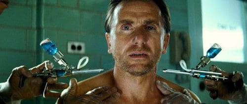 A man with a concerned expression sits in a clinical setting, while two gloved hands hold syringes poised toward his shoulders. The background features a tiled wall and medical equipment.