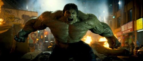 The Hulk, a massive humanoid figure with green skin and bulging muscles, roaring in an urban setting filled with chaos. Buildings are illuminated by flames in the background, and debris is scattered around the street.