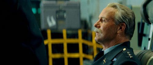 A man with gray hair and a mustache wearing a military uniform, sitting in a command area. His expression is calm as he looks off to the side, with a background featuring a blurred yellow safety barrier.