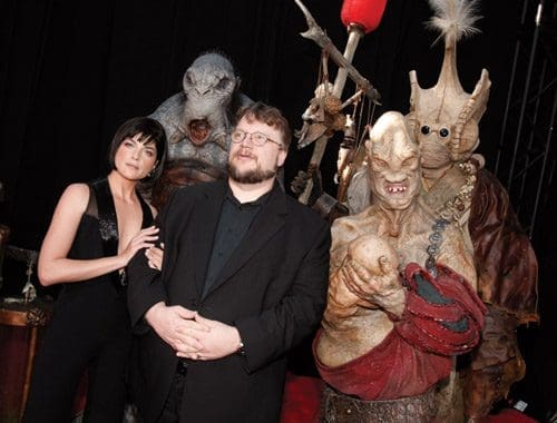 A woman in a black outfit stands next to a man in glasses and a black suit, both smiling. Behind them are two fantastical creatures in elaborate costumes, showcasing dramatic masks and details, against a dark background.
