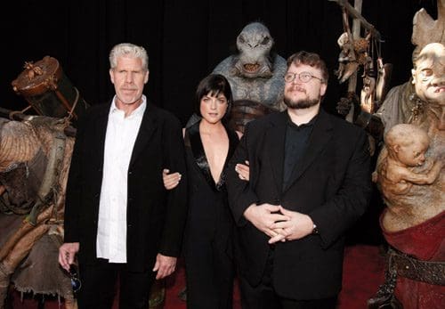 Three individuals pose on a red carpet at an event, surrounded by large, fantastical creature puppets. The first person, with gray hair and a black suit, stands on the left. The woman in a black outfit is center, while the man in glasses and a black jacket is on the right.