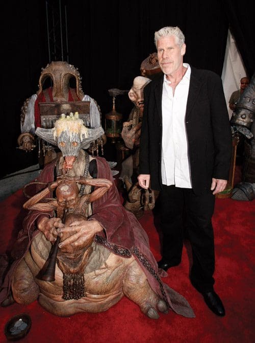 A man standing on a red carpet next to a larger-than-life, intricate creature sculpture with exaggerated features, surrounded by other artistic figures. The sculpture is detailed, showcasing various textures and designs.