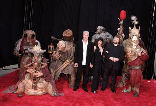 A group of people posing on a red carpet with various fantasy creature costumes. The central figures are dressed in black, standing beside intricately designed creature characters, adding a whimsical and imaginative atmosphere.