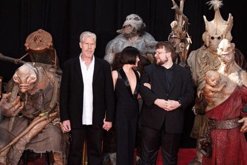 Three individuals pose in front of elaborate creature costumes on a red carpet, showcasing intricate designs and details. The backdrop is dark, highlighting the unique figures, including a creature with a large head and various fantastical elements.