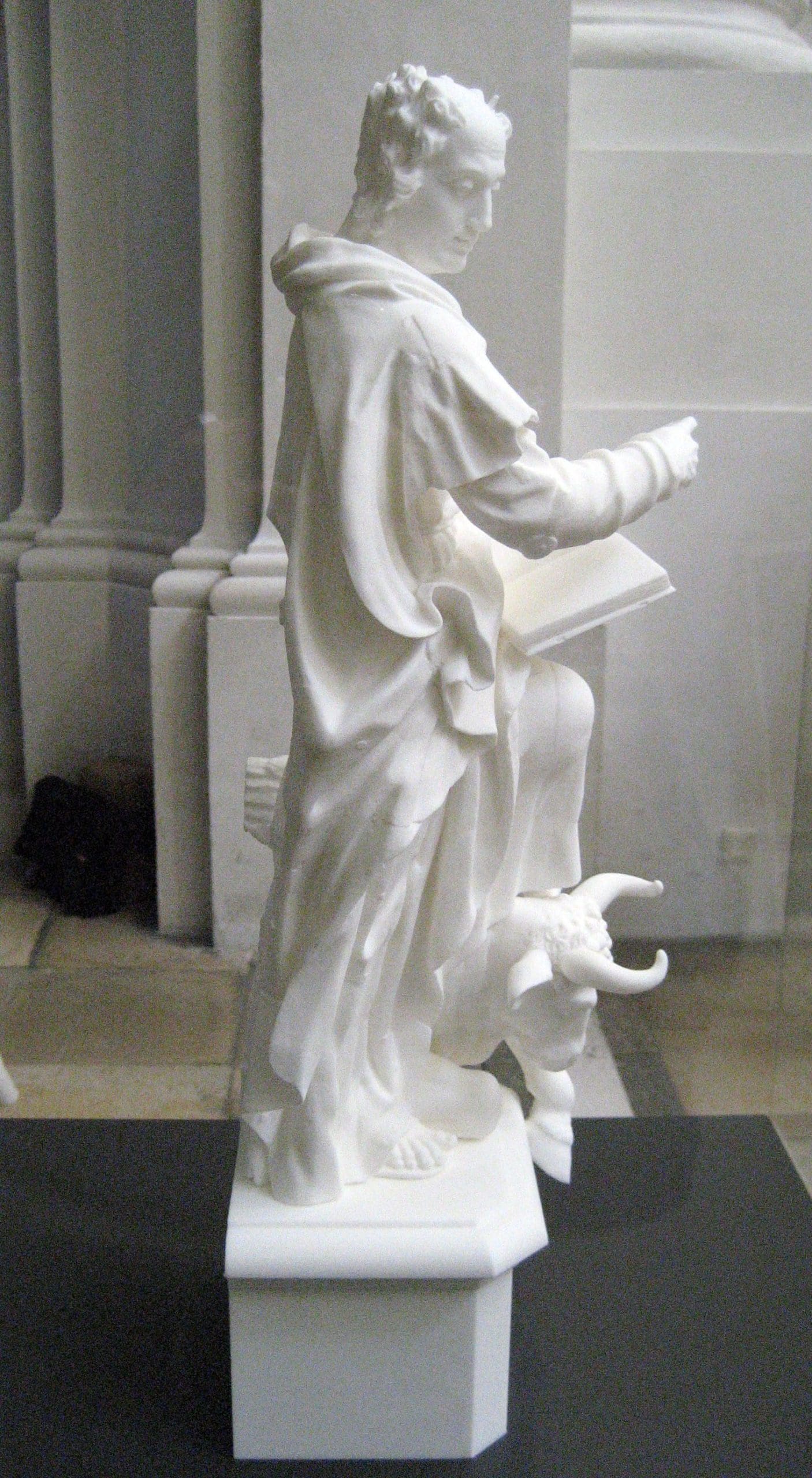 White sculpture of a standing figure holding a book, with a creature at the base.