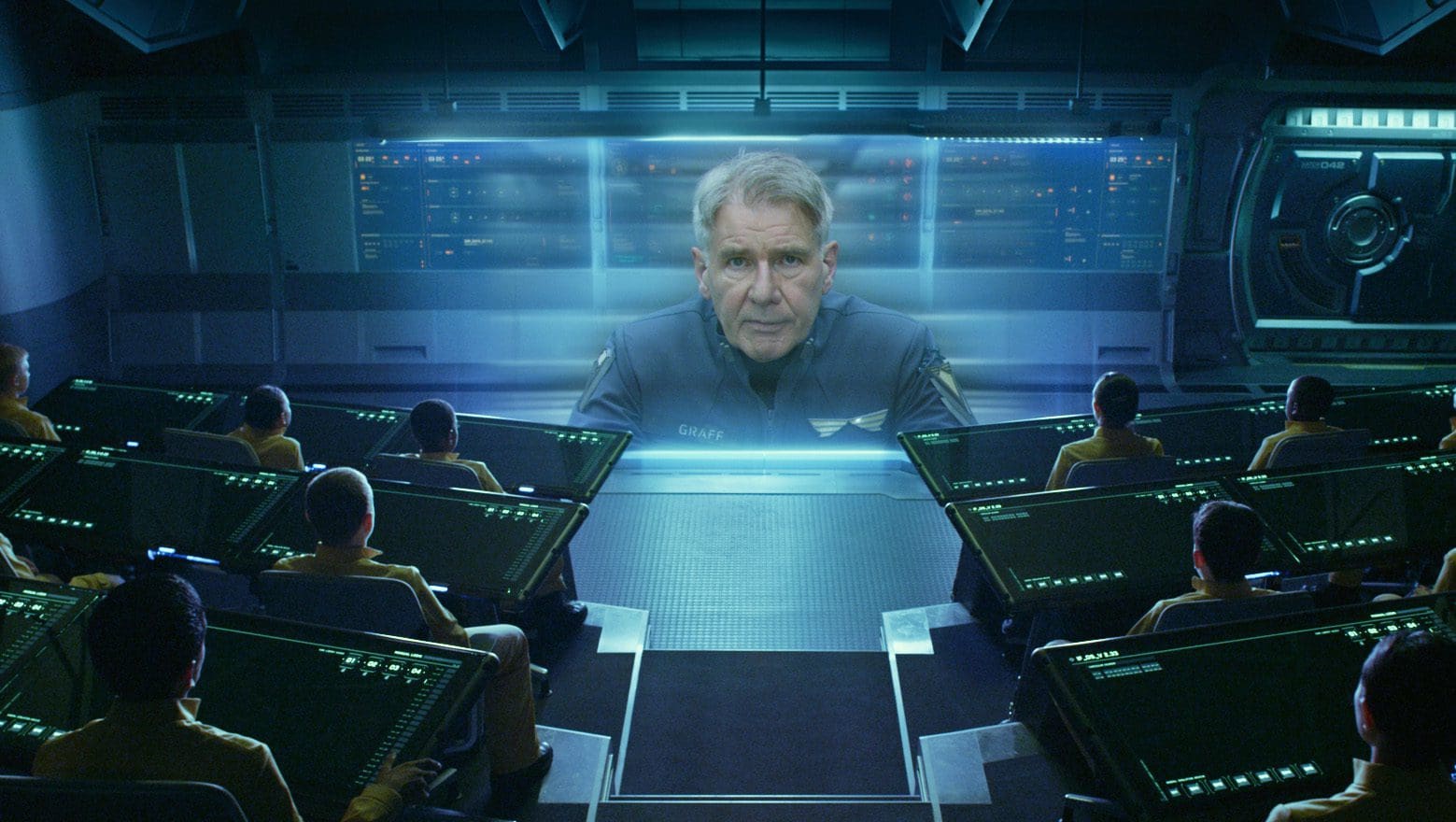 Man speaking in hologram to crew in futuristic control room.