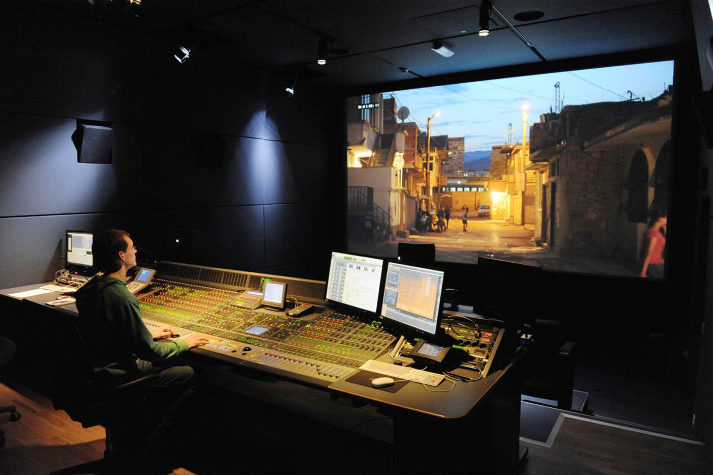 Audio engineer working at a sound mixing console with a city scene displayed on a large screen behind.
