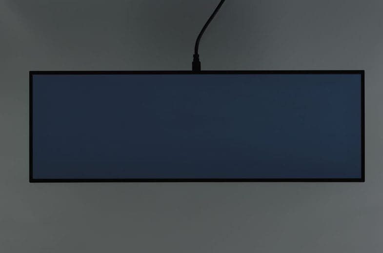 A slim black rectangular monitor with a dark screen hanging against a grey background.
