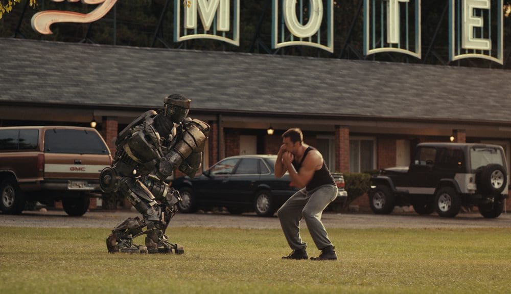 A man and a robot facing off in a grassy area near a motel.
