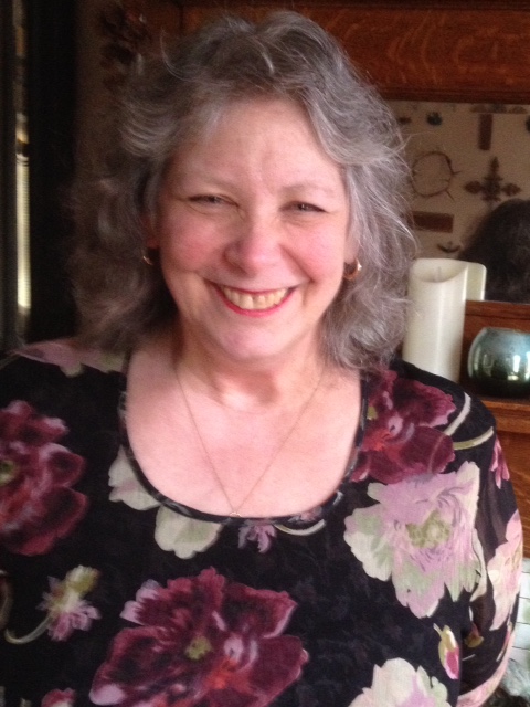 An older white woman with long gray hair. She is wearing a black top with flowers.