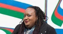Cropped image of Imani standing on her crutches in a blue dress and jacket in front of a blue red and green mural wall. Photo credit: Madasyn Andrews
