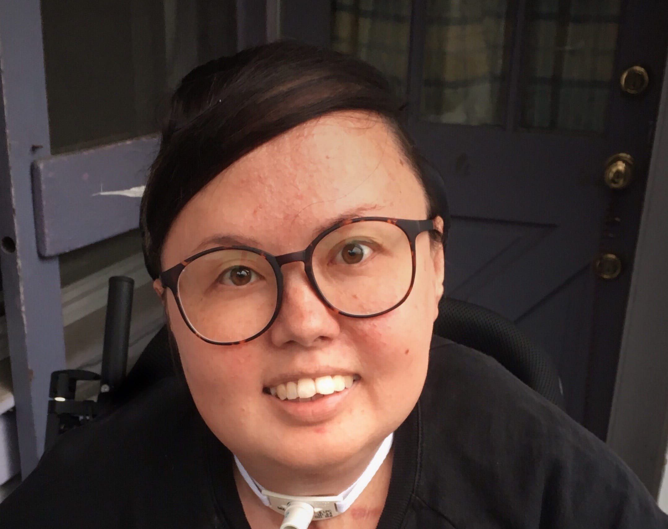 A mixed race Korean and white queer person smiles head on at the camera. She has big glasses and is wearing a crewneck. Her trach and wheelchair can be seen.