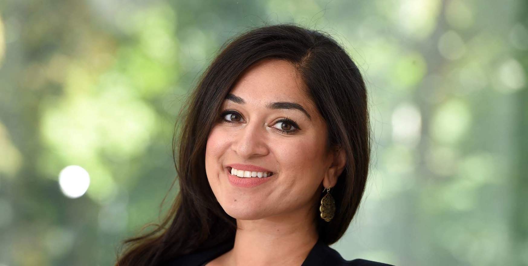 Jaipreet Virdi, a South Asian woman with dark hair, smiles at the camera. Her head is slightly turned. She is wearing a black blazer and dark top. There are some trees in the blurred background.