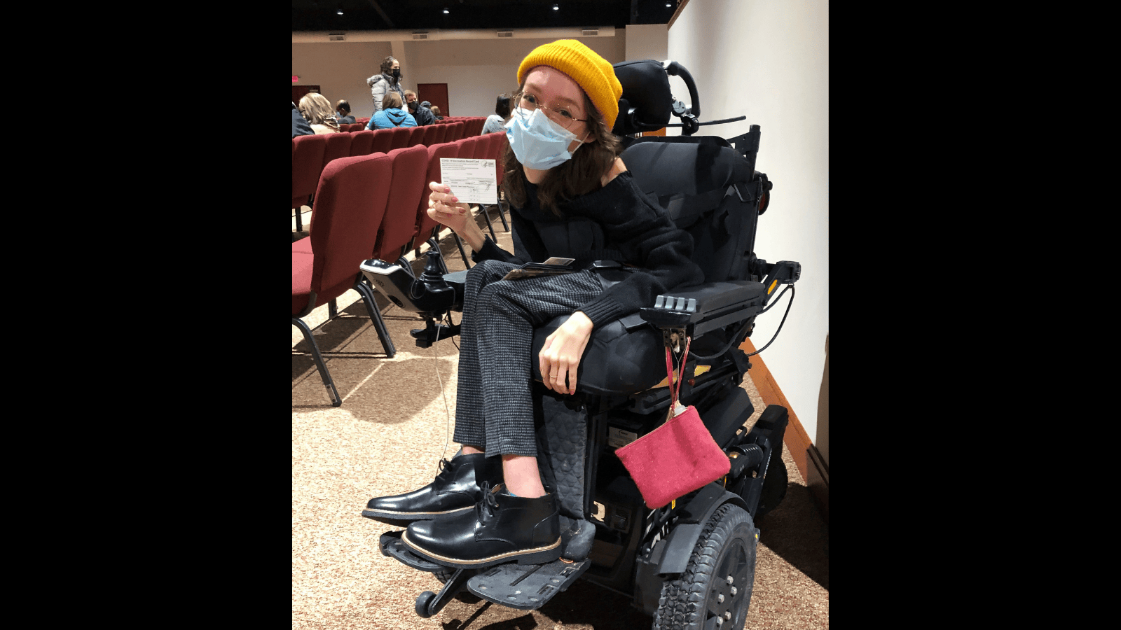 Graphic with a black background and a photo of Emily Ackerman, a white woman in a black power wheelchair is holding up her COVID vaccine card. She’s wearing a black sweater and boots with grey checked pants, a yellow beanie, and a blue medical mask. 