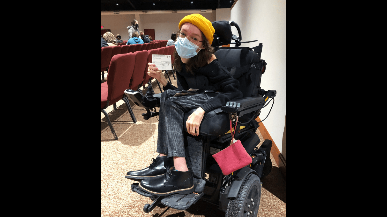 Graphic with a black background and a photo of Emily Ackerman, a white woman in a black power wheelchair is holding up her COVID vaccine card. She’s wearing a black sweater and boots with grey checked pants, a yellow beanie, and a blue medical mask. 