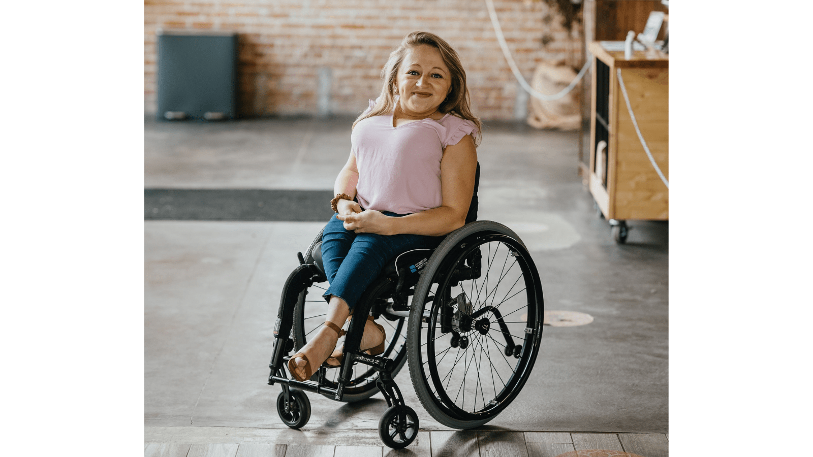 A white woman, Emily, with blonde, shoulder-length hair sits in a black wheelchair. She is short of stature. She is wearing a pink short-sleeved shirt and blue jeans with brown wedge heels. The ground is partially cement, partially wood. There is a brick wall behind her.