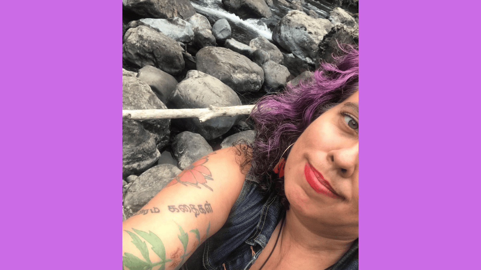 Graphic with a light purple background with a photo of a middle aged mixed race Sri Lankan, Irish and Romani femme, diagonially, in front of a wild river with lots of rocks. Leah has violet, brown and silver curly hair, peachy tan sand colored skin and red lipstick (Stila Stay All Day lipstick in Beso, aka "the lipstick AOC wears.") Their clothes are mostly not visible except for a blue denim vest. You can see their sleeve of tattoos, including cosmos flowers, a motherwort plant and letters in Tamil. They are smiling with bashful pride and satisfaction."