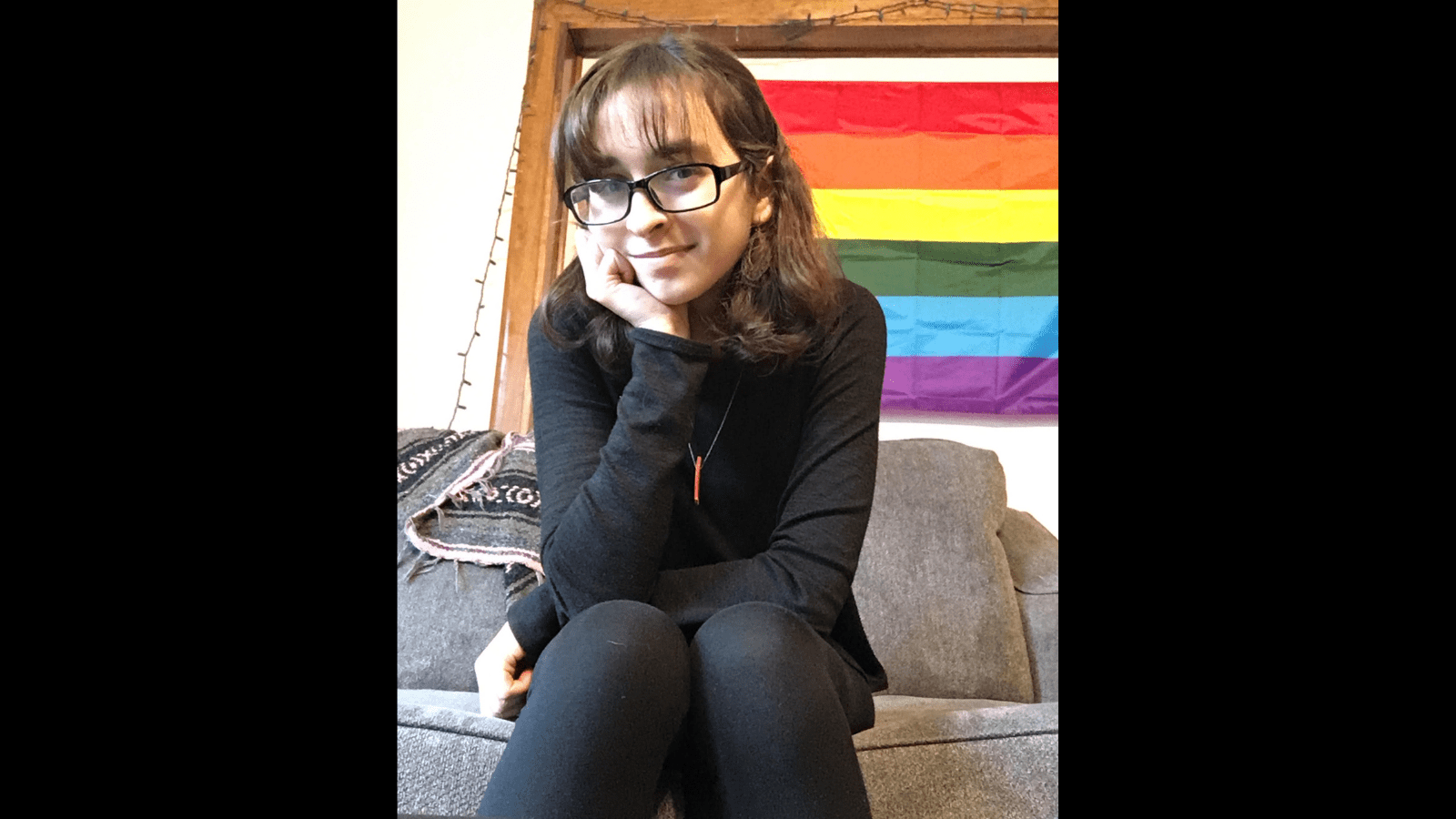 Graphic with a black background featuring a photo of Rachel, a white femme-presenting person sits in front of a pride flag. She has brown shoulder-length hair, and is wearing bronze earrings, black clothing, and black rimmed glasses. A little red piece of wood dangles from a thin silver necklace chain around her neck. She is sitting on a grey couch and rests her chin in one hand with one elbow on her knee. She grins slightly at the camera.