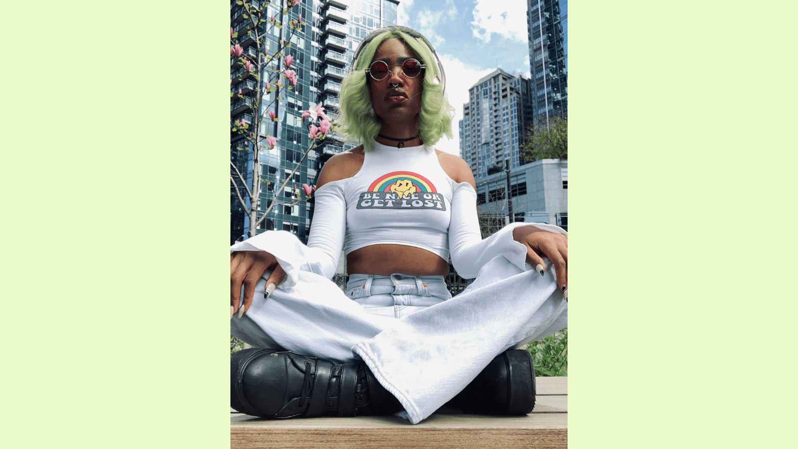 Image of Faati, a nonbinary light brown skinned Black Native, sitting cross legged in front of a cherry blossom tree and multiple high rise buildings. They have on acid washed light blue bell-bottom jeans, a white crop topped long sleeved shirt and black platform boots. They are sitting cross-legged with their hands on their knees. Their fingernails are a long almond acrylic style with nude coloring and black and white designs on top. They have green straight 70s styled hair with the ends bumped. They are wearing circle 70s style red lens sunglasses.