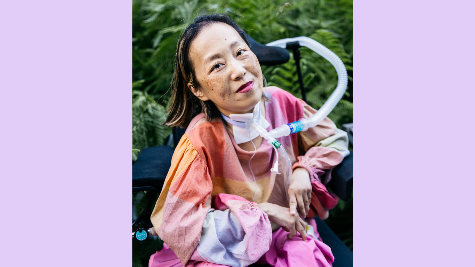 Graphic with a purple background. In the center is a photo of an Asian American woman in a wheelchair with a tracheostomy at her neck that has a tube connected to her ventilator. Her head is tilted to the left side, she is smiling, and she is wearing a magenta lip color. Her shirt is plaid with pale pink and other pastel colors and she is wearing pink pants. Behind her is a lot of greenery from bushes and trees at a park. Photo by @allisonbusch_photography