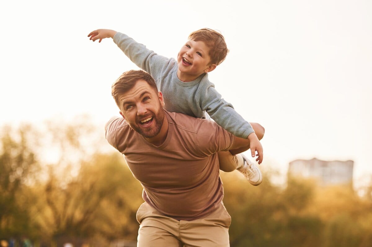 A man smiles while giving a piggyback ride to a young boy outdoors. The boy, dressed in light blue, spreads his arms wide as if flying, both appearing happy. The background shows soft-focus trees and a light sky with a distant building. Read more at Divorce Squad (divorcesquad.com)