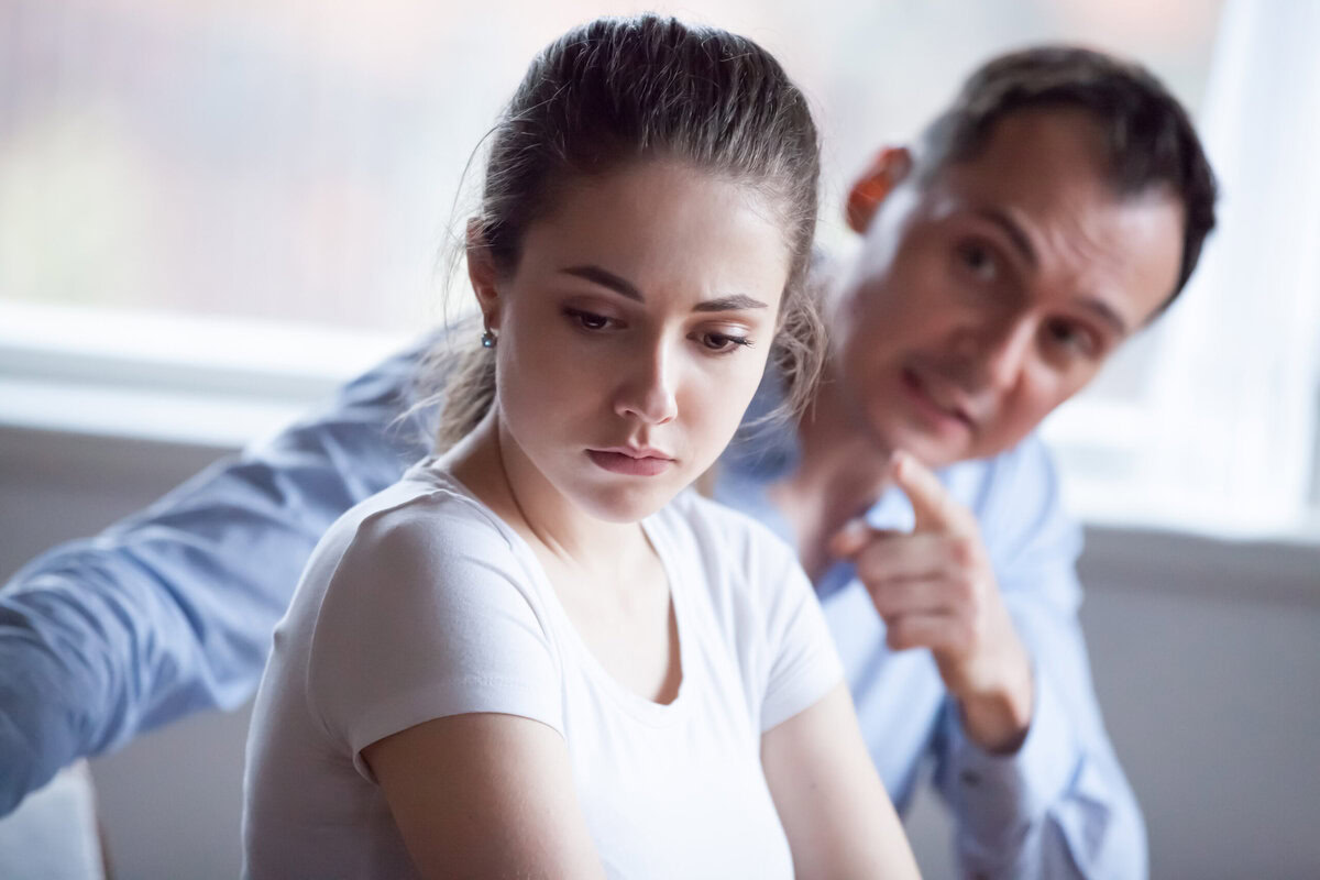A young woman with long brown hair in a ponytail looks down sadly, avoiding eye contact. Behind her, a man in a blue shirt gestures sternly—capturing the tense mood often seen when divorcing a narcissist. Read more at Divorce Squad (divorcesquad.com)