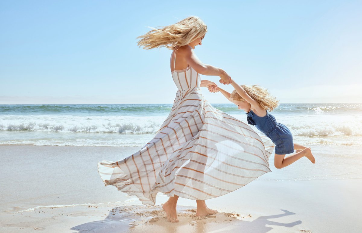 A woman in a white striped dress spins a young girl in a blue dress by the arms on a sunny beach, savoring vacation time together. Both have windswept blonde hair as ocean waves roll in—a cherished moment for moms navigating custody agreements. Read more at Divorce Squad (divorcesquad.com)
