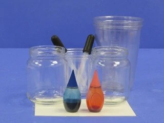 A collection of glass jars and cups with two bottles of blue and red food coloring in the foreground, set against a blue background.