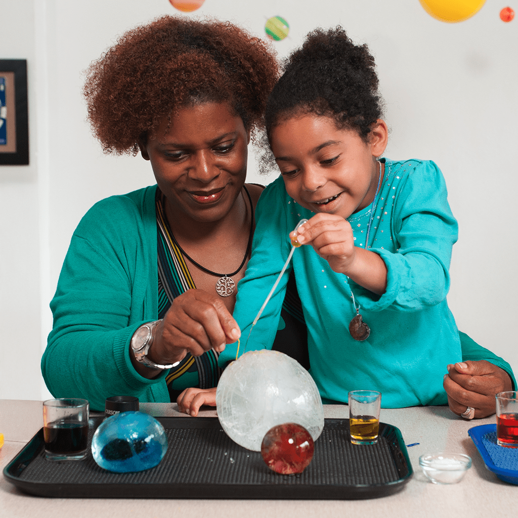 An adult and a young child working together on a science experiment, using pipettes to drop colored liquid onto large spheres of ice.