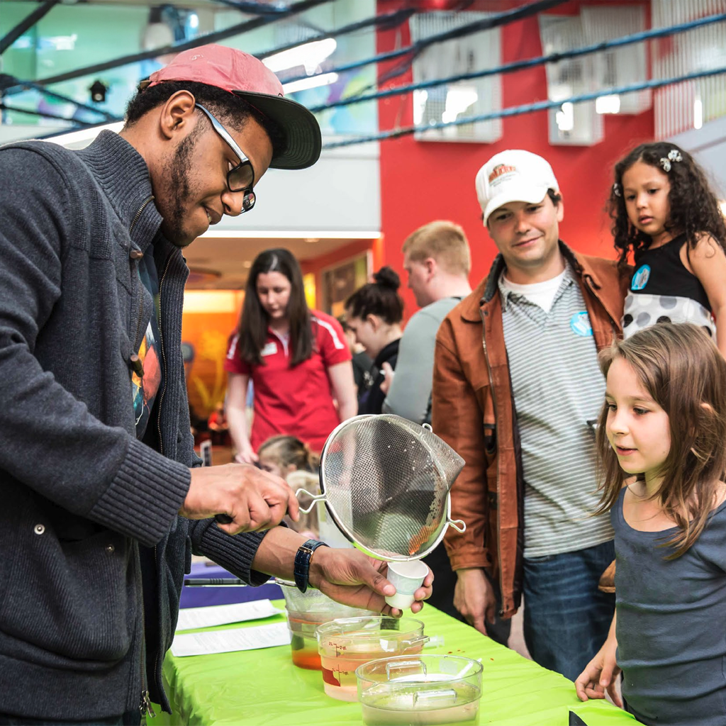 A facilitator in a grey sweater and cap demonstrates a hands-on science activity, pouring liquid through a strainer while a young girl and her family watch intently at a table.