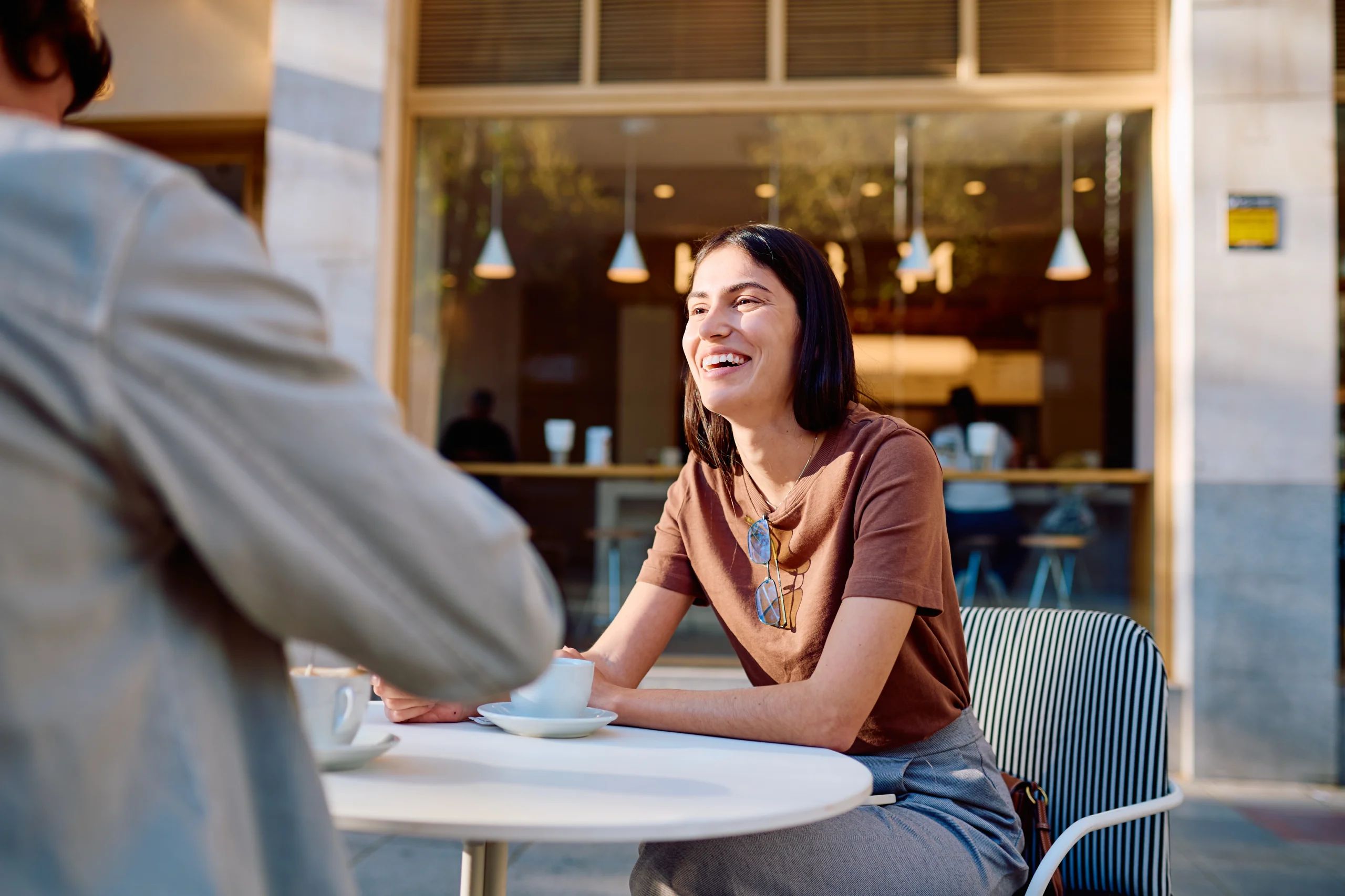 A smiling young woman sitting at a round outdoor cafe table having coffee and conversation with a companion.