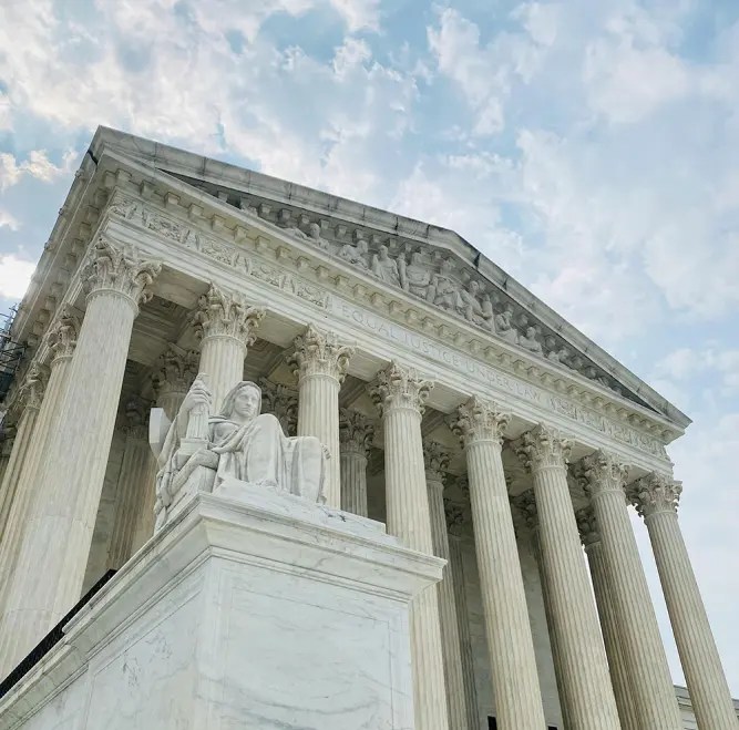 Một góc nhìn từ dưới lên của tòa nhà Tòa án Tối cao Hoa Kỳ tại Washington D.C., nổi bật với mặt tiền có các cột kiểu Corinthian và một tác phẩm điêu khắc hình người đang ngồi ở phía trước.