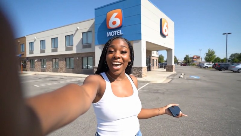 Content creator surprised by bidet in fancy Motel 6 bathroom A smiling woman standing in front of a Motel 6, holding a phone and dressed in a white tank top and jeans, with the motel building and parking lot visible in the background.