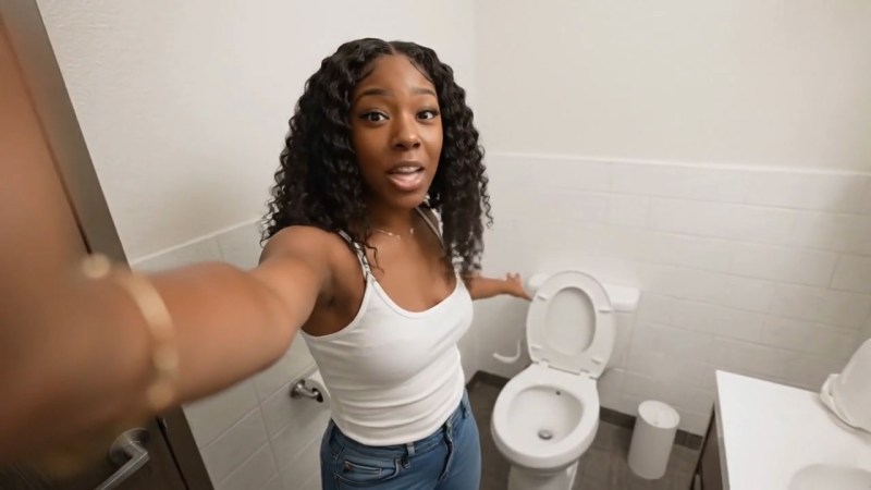 A woman standing in front of a bidet in a bathroom, pointing towards a toilet while smiling, showcasing her surprise and excitement.