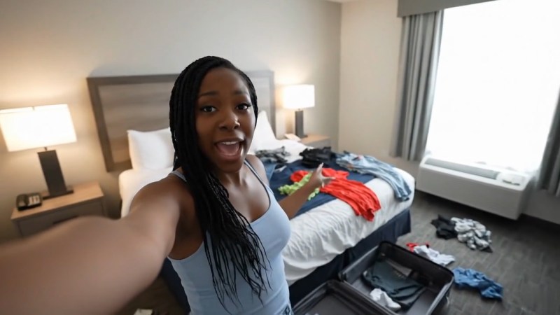 A person in a hotel room, standing next to a messy bed with clothes scattered around, smiling and gesturing enthusiastically.