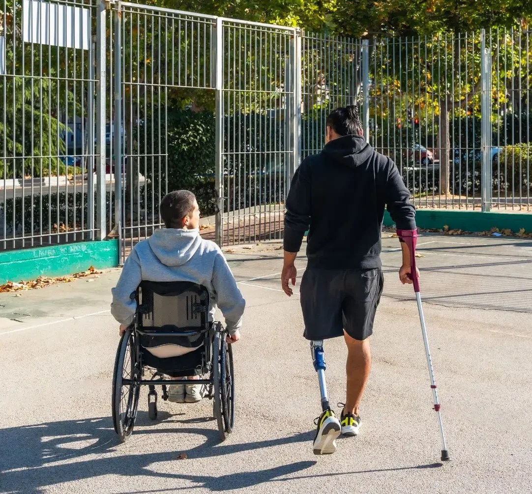 Two people outside on a basketball court. The person on the left is using a wheelchair, the person on the right has a cane and a prosthetic leg.