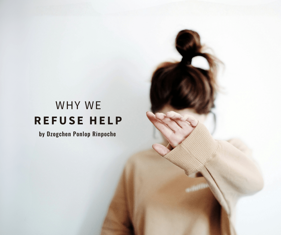 Figure is shown standing against a light gray wall. They are pictured from the waist up, wearing a loose-fitting tan sweater and hair pulled up in brown topknot. Their left arm is raised, with hand up, covering their face.